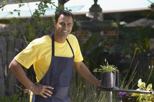 A man wearing a yellow T-shirt and a dark apron is standing outdoors in a garden, smiling at the camera while holding a small black plastic container with a young potted plant. The garden features a well-maintained lawn with dense, green grass in the foreground, and behind him, there are various plants, shrubs, and a decorative stone fountain situated among garden beds. To the right, there's a small, circular metal table supporting the plant pot. The background shows a wooden fence, some tall plants, and a structure with a green canopy, all under natural light with clear weather conditions. The scene exemplifies typical gardening activities, such as planting or transplanting, which are relevant to outdoor garden maintenance and landscaping services provided by companies like Hedge Trimming Soho in the local area near Soho or central London. The overall environment indicates a neatly landscaped backyard designed for outdoor enjoyment and horticultural care.