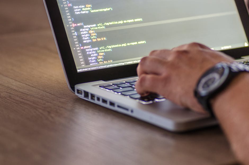 A close-up image of a person's hand typing on a silver laptop keyboard placed on a wooden desk. The screen displays lines of colorful code, indicating programming or web development work. The person's