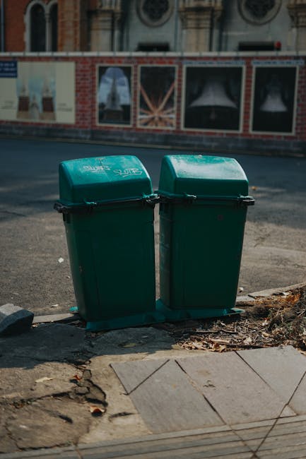Two green garden waste bins with black handles and lids, positioned side by side on the edge of a paved sidewalk near a curb. The bins are made of plastic and are slightly weathered, with minor scratc